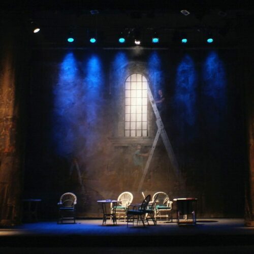 A stage set with chairs and tables under blue spotlights and a painted backdrop